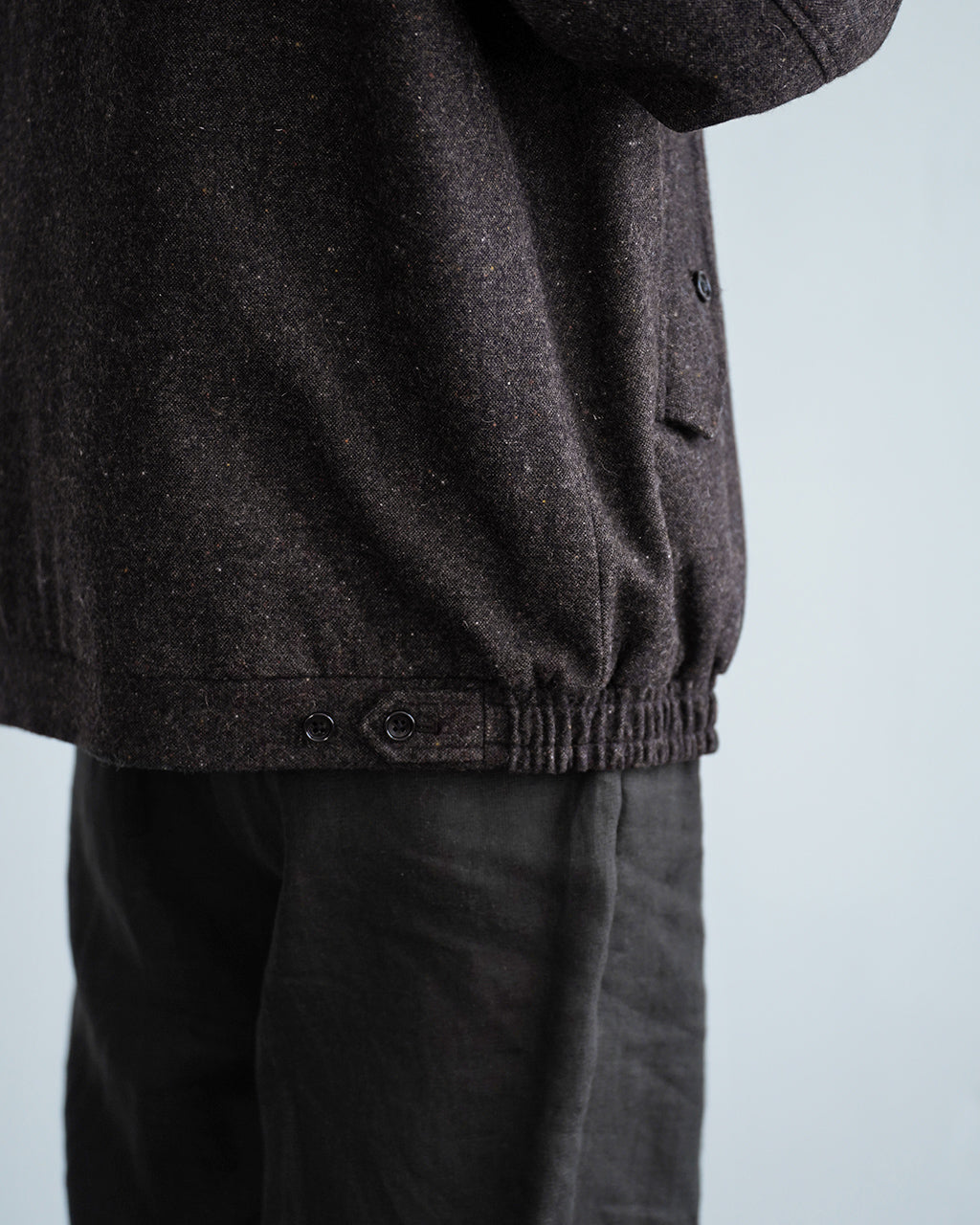 黒系のヘリンボーン風生地のハリ感ある Harrington ジャケットの裾部分。絞りのあるウエストとボタン留めアジャスターが控えめに見えるディテール画像