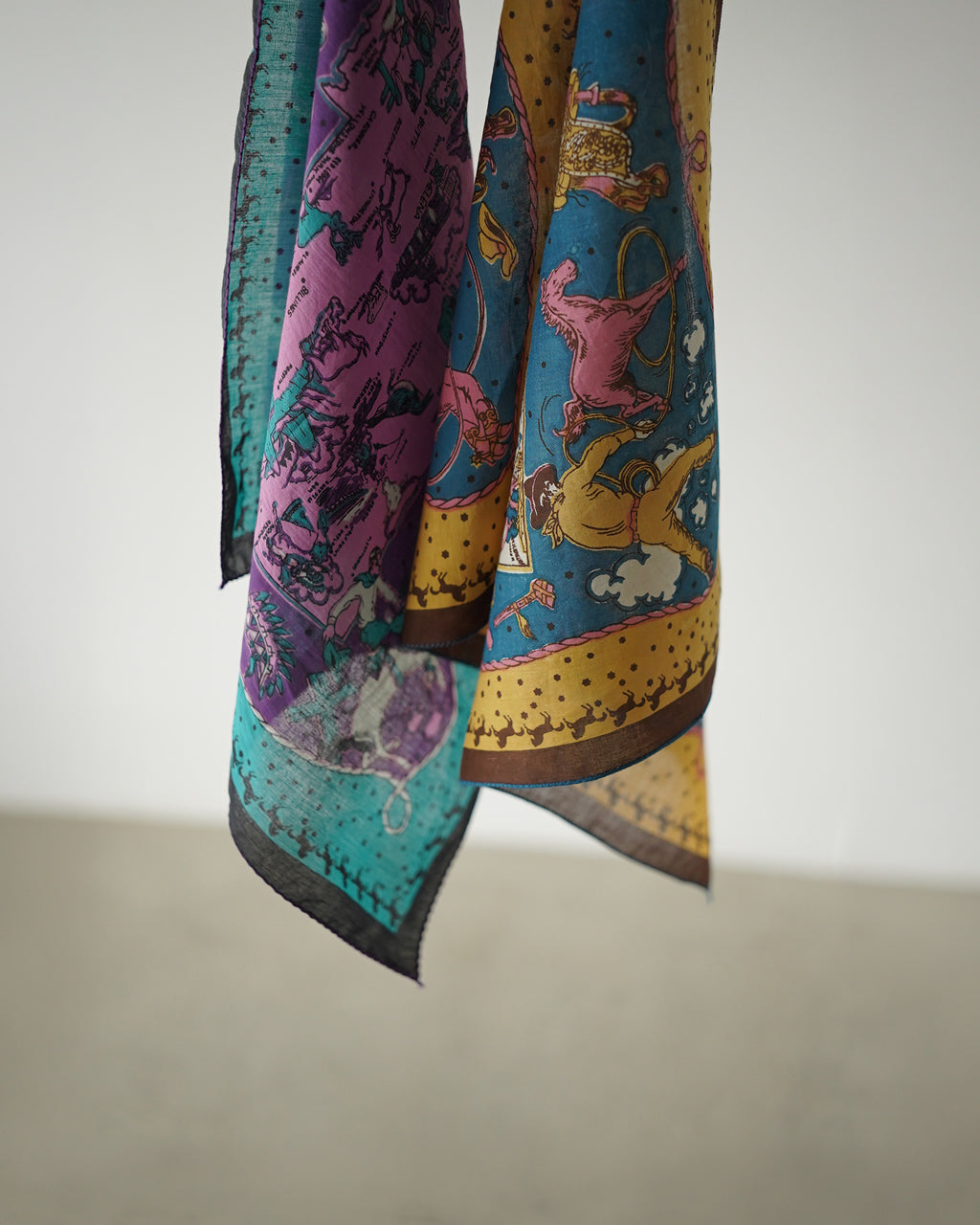 Montana Bandana 50×50cmのカラー柄が映える生地が垂れ下がる姿。緑・紫・青の色ブロックに、装飾的なモチーフが散りばめられた大判スカーフ風デザイン。