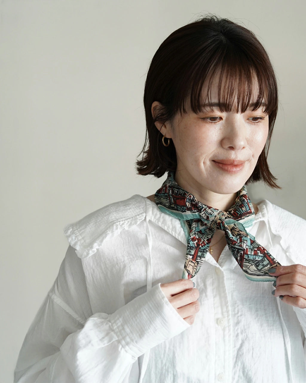 白いシャツにバンダナを結んだ女性の正面寄りのアップ。柄の細部が見えるカラーのストールで、首元のアクセサリーも映るスタイル