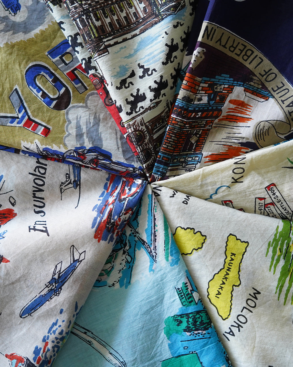 色とりどりの地図風デザインが広がるSCARF、64×64cmの正方形スカーフ。柄は旅の名所が散りばめられ、折りたたまれた状態でカラフルに配置されている。