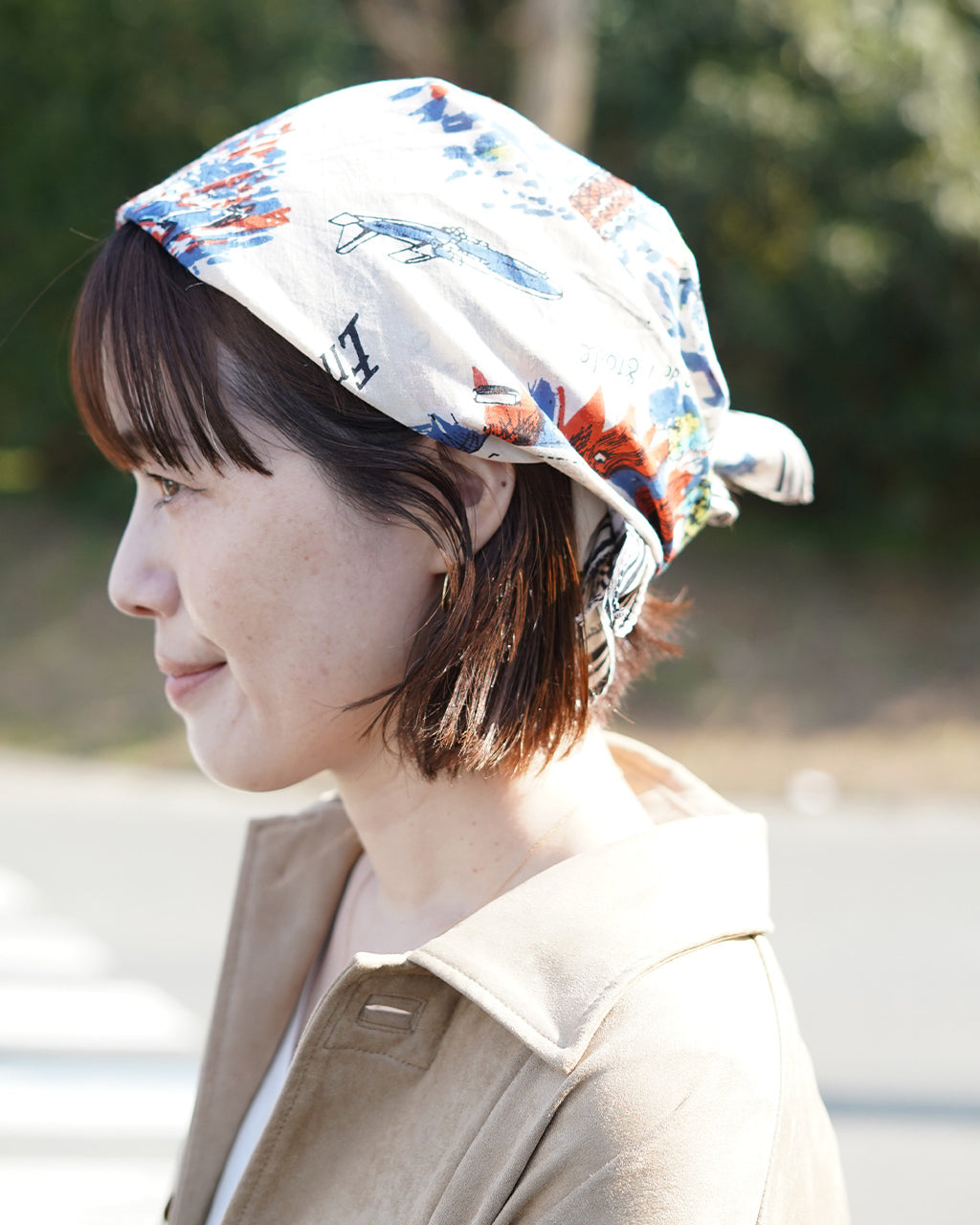 白地に絵柄が散らばるスカーフを頭に巻いた女性の横顔。やわらかな光で自然な雰囲気、首元にはベージュ系のコートが映えるシーンです。
