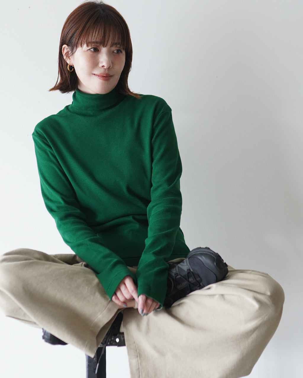 緑のタートルネック長袖シャツを着る女性の正面やや斜めからの姿。ベージュのパンツと黒系の靴を合わせ、リブ編みと首元のタートルデザインが際立つコーディネート。