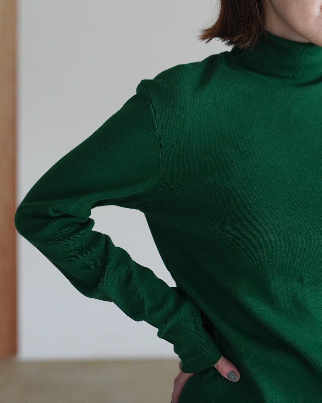 深緑のタートルネック長袖Tシャツのアップ。細いリブ編みが施され、袖口と首元の繊細な縫製が見える着用イメージ。