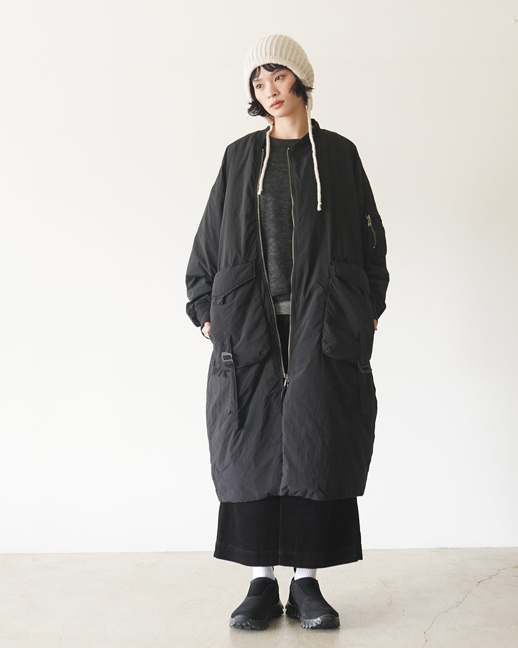 ブラック系のロングMA-1ダウンコートを着用した女性が全身で写る画像。前面ジップ開閉と大きめポケット、ドロップショルダーのボリューム感が特徴的なデザイン。ニット帽とスニーカーを合わせたカジュアルなスタイル。