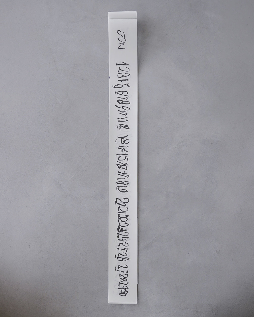 縦長の短冊型カレンダーの表紙を示す画像。紙を白地に黒の手書き風数字が並ぶデザインで、壁に掛けるストラップが付属する点が読み取れる。