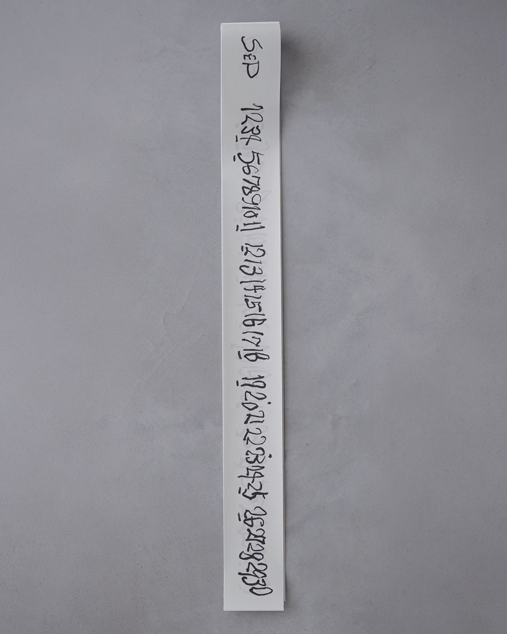 縦長の白い紙に黒い手書き文字が連なる、中央に立て掛けられた長冊タイプのカレンダー。グレーの壁を背景に撮影され、表紙のような札が先端に見える。