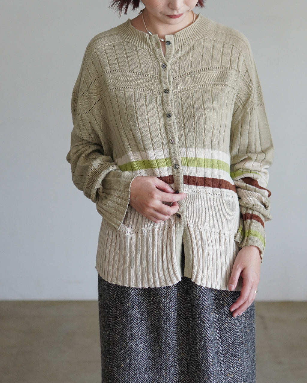 ベージュ系のコットンニット cardigan。ボタンで前開き、袖と裾にリブとアイレット柄、横ストライプ風の配色ラインが特徴。