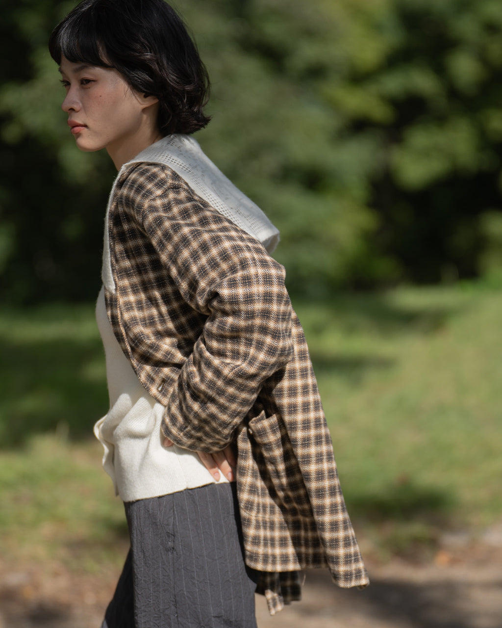 Brown-Checkのジャケットを羽織る女性の横顔と肩のラインが写る野外写真。チェック柄の布と白いインナーがコントラストを作り、風合いが伝わるディテール写真です。