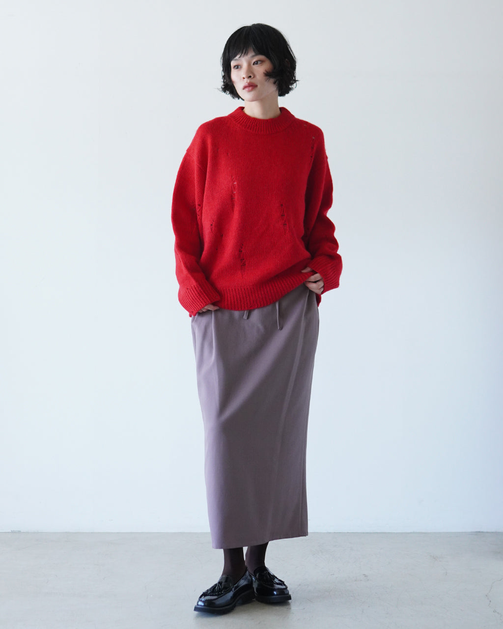 赤いざっくりニットを着用した女性の全身ショット。モーブ系スカートと黒のシューズで大人っぽく着こなし、腰周りは緩やかなドレープ感とゆとりのあるペンシルラインが特徴です。