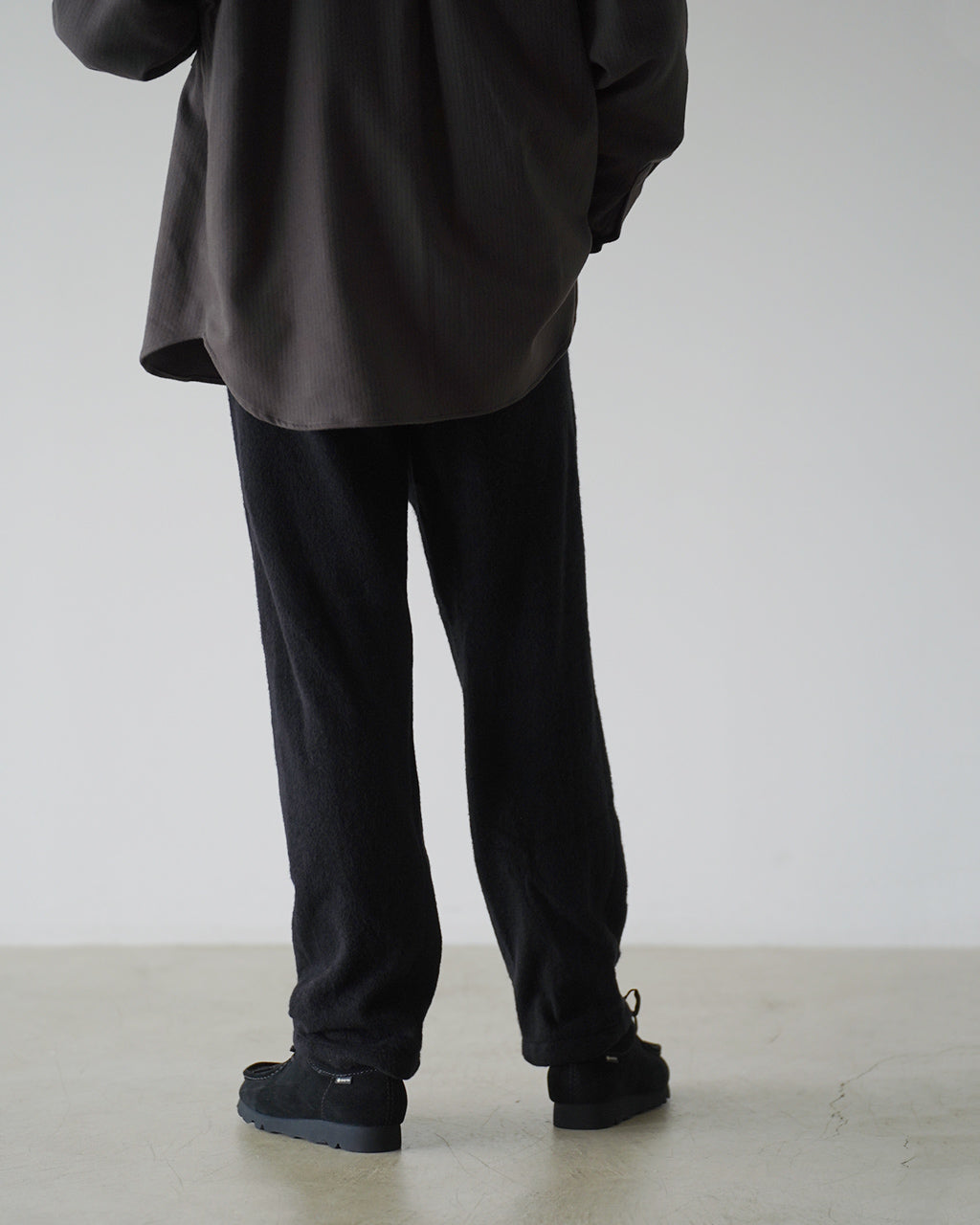 背面寄りの着用イメージ。黒系のSilk Wool Fleece Pantsとゆるく落ちる裾のシルエットが分かる写真。腰回りはややゆとりがあり、裾へ向かって細くなるラインを確認できる。