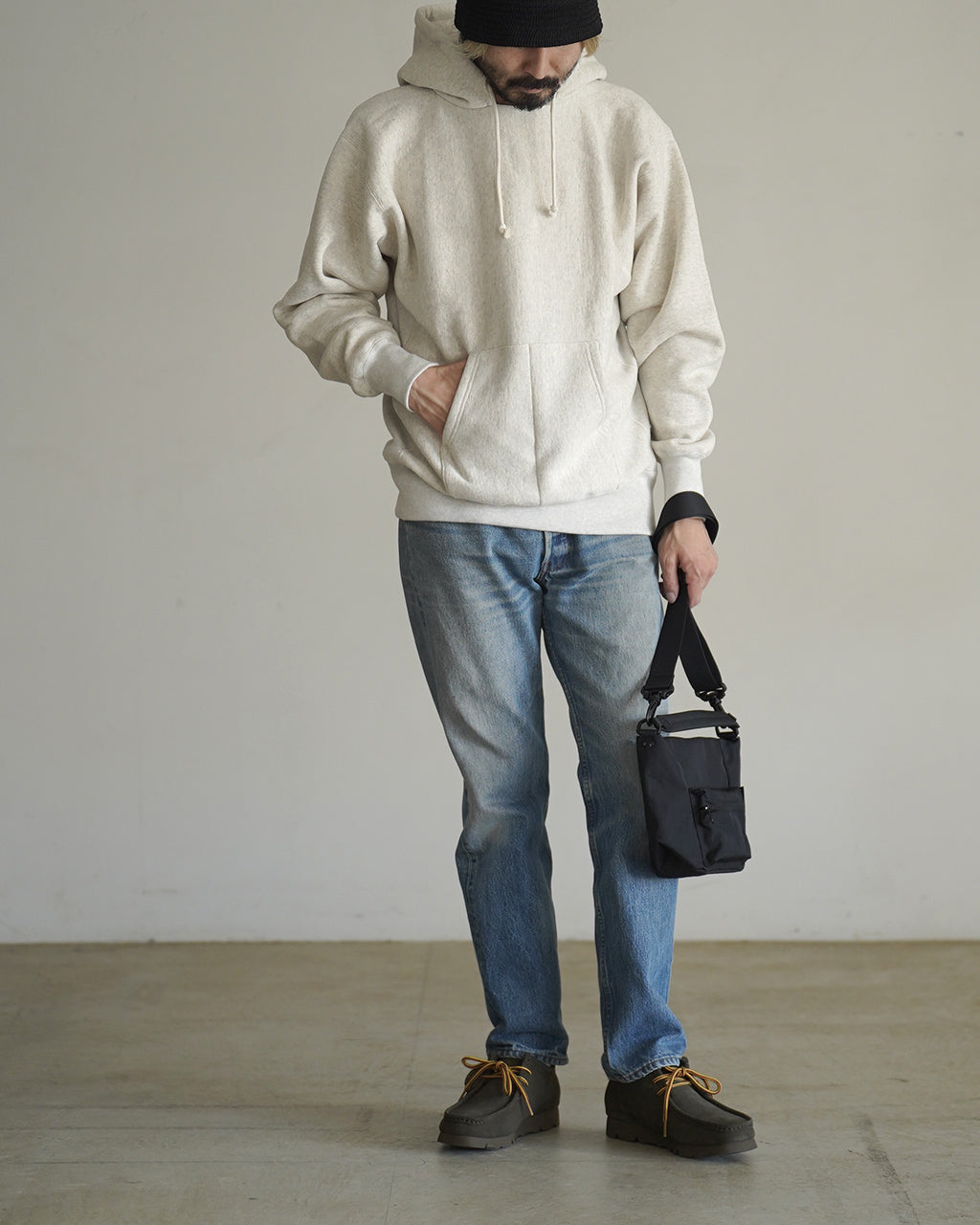 白系のパーカーを着た男性が2WAYショルダーバッグを手に持つ様子。カジュアルな outfit に調和するコンパクトなデザインが分かる前面アングル。