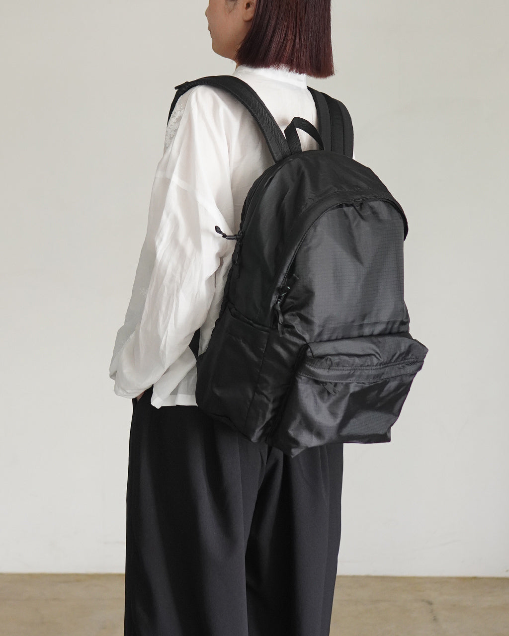 黒色のバックパックを背負ったモデルの後ろ姿。白いシャツと黒系パンツのコーディネートで、背面の大きなリュックが目を引く写真です。