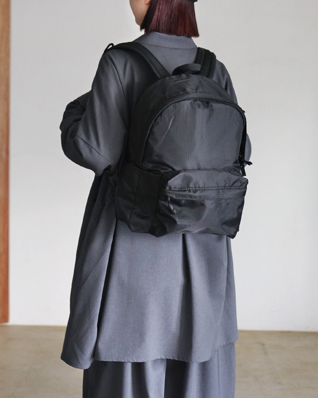 LC by LOWERCASEのDAYPACK LITE 16Lを背負った女性の後ろ姿。黒系のバックパックとグレーの長袖コートが映える着用イメージ。