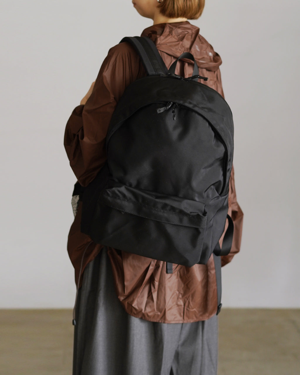 ブラックの DAYPACK Msize を背負うモデルの後ろ姿。丸みを帯びた大きなリュック本体と背面ストラップが際立つ、シックな印象のバッグ写真。