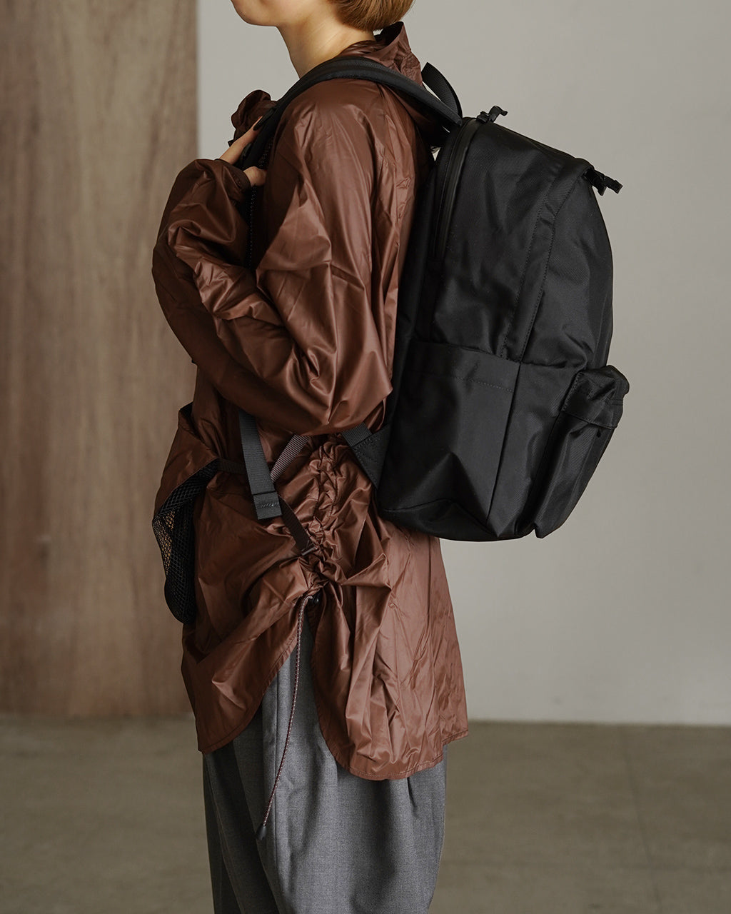 ブラックの DAYPACK Msize を背負うモデルの横背面ショット。肩ストラップとサイドポケット、前後のシルエットが分かる構図。
