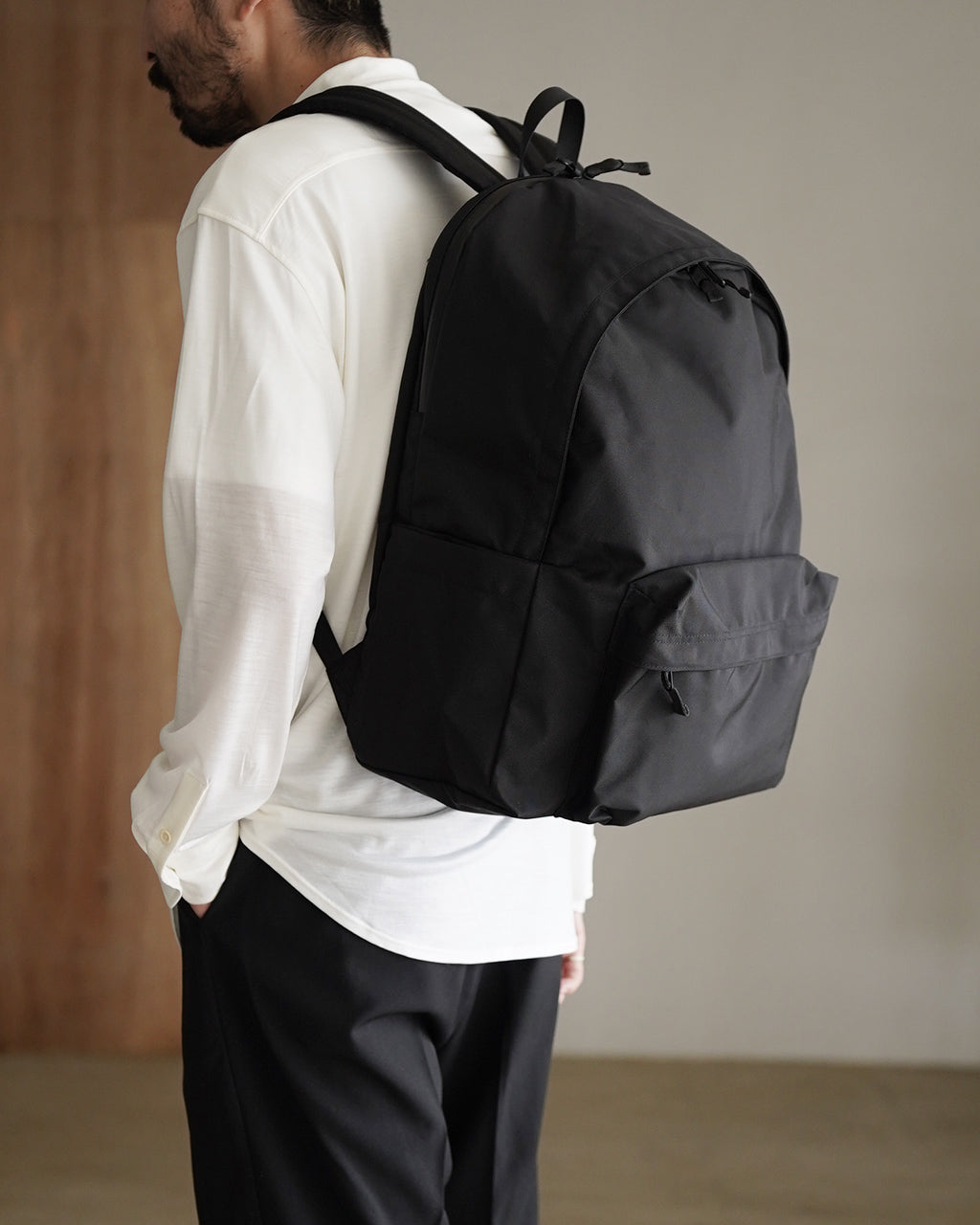 ブラックの DAYPACK Lsize を背中に背負った男性の横姿。シンプルなデザインで、背面とサイドポケットが見える。