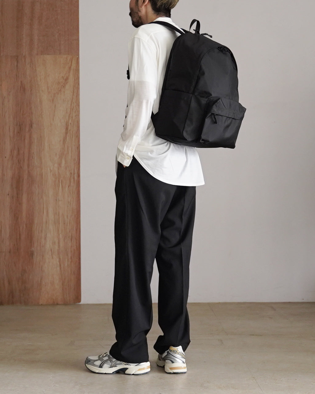 黒の DAYPACK Lsize を背負う男性の後ろ姿。大きめのバックパックは前方のポケットが目立ち、シャツとパンツのモノトーンコーデと調和している