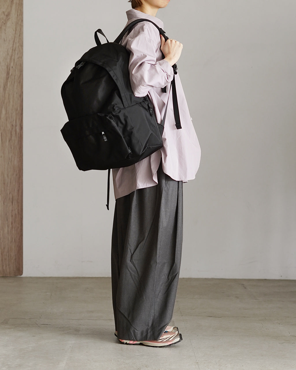 黒色の DAYPACK Lサイズを背負う女性の横からのシルエット。大きめのバックパックと薄いピンクのシャツ、グレーのパンツが調和したコーディネートが映える写真。