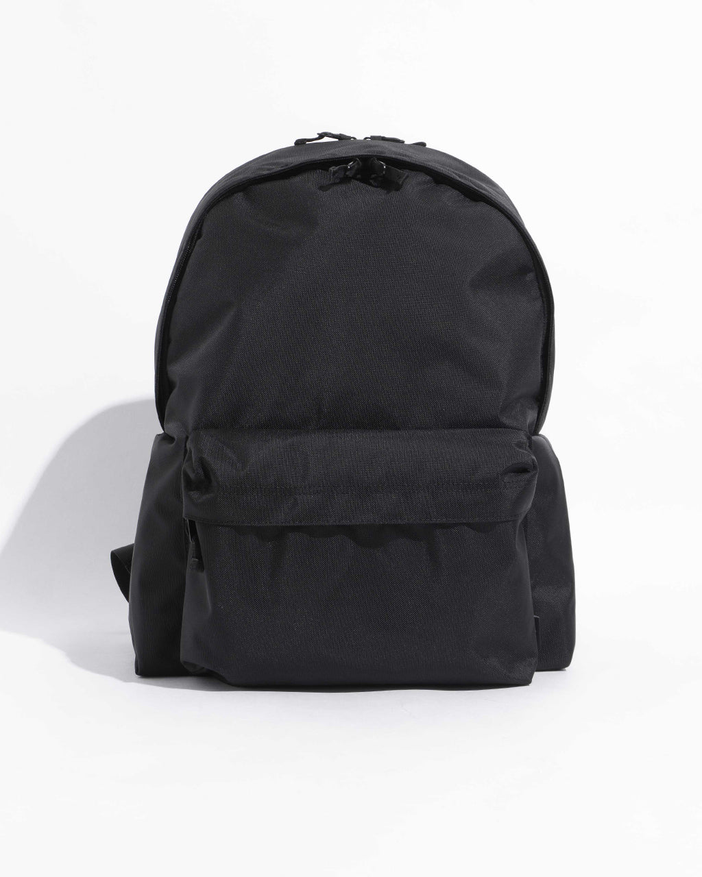 DAYPACK Lサイズは無地の黒系ナイロン地で、前後ろの丸みを帯びたシルエット。背面部に大きめのフロントポケットとサイドポケットが特徴的なバックパックです。