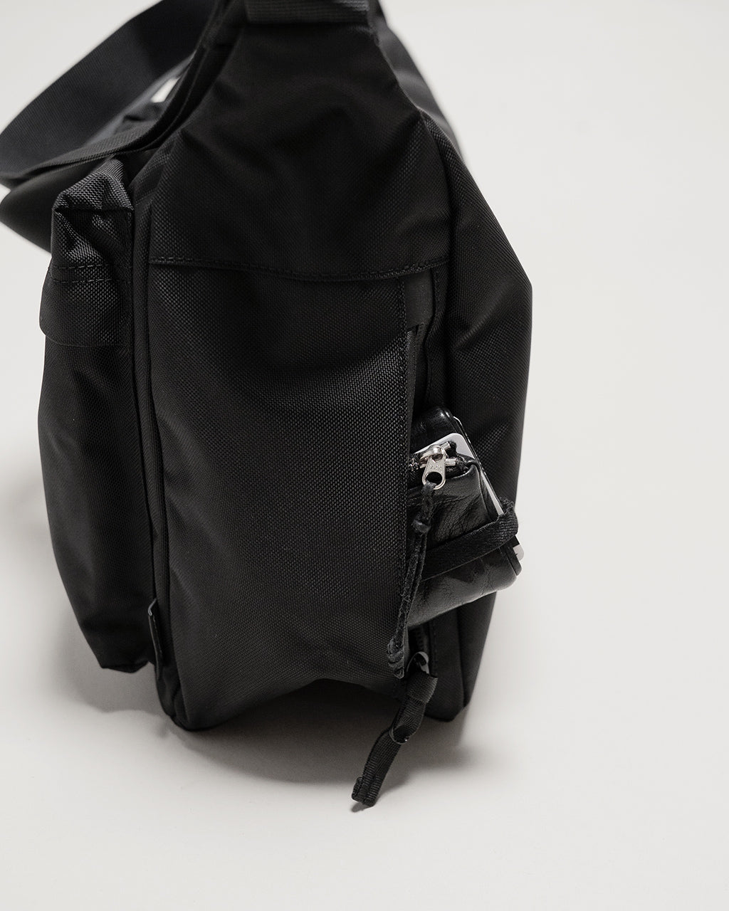 黒のショルダーバッグ。側面のポケットと金具付きのジッパーが見える、ナイロン製のミニマルなデザイン。容量と丈夫さが伝わる構造です。
