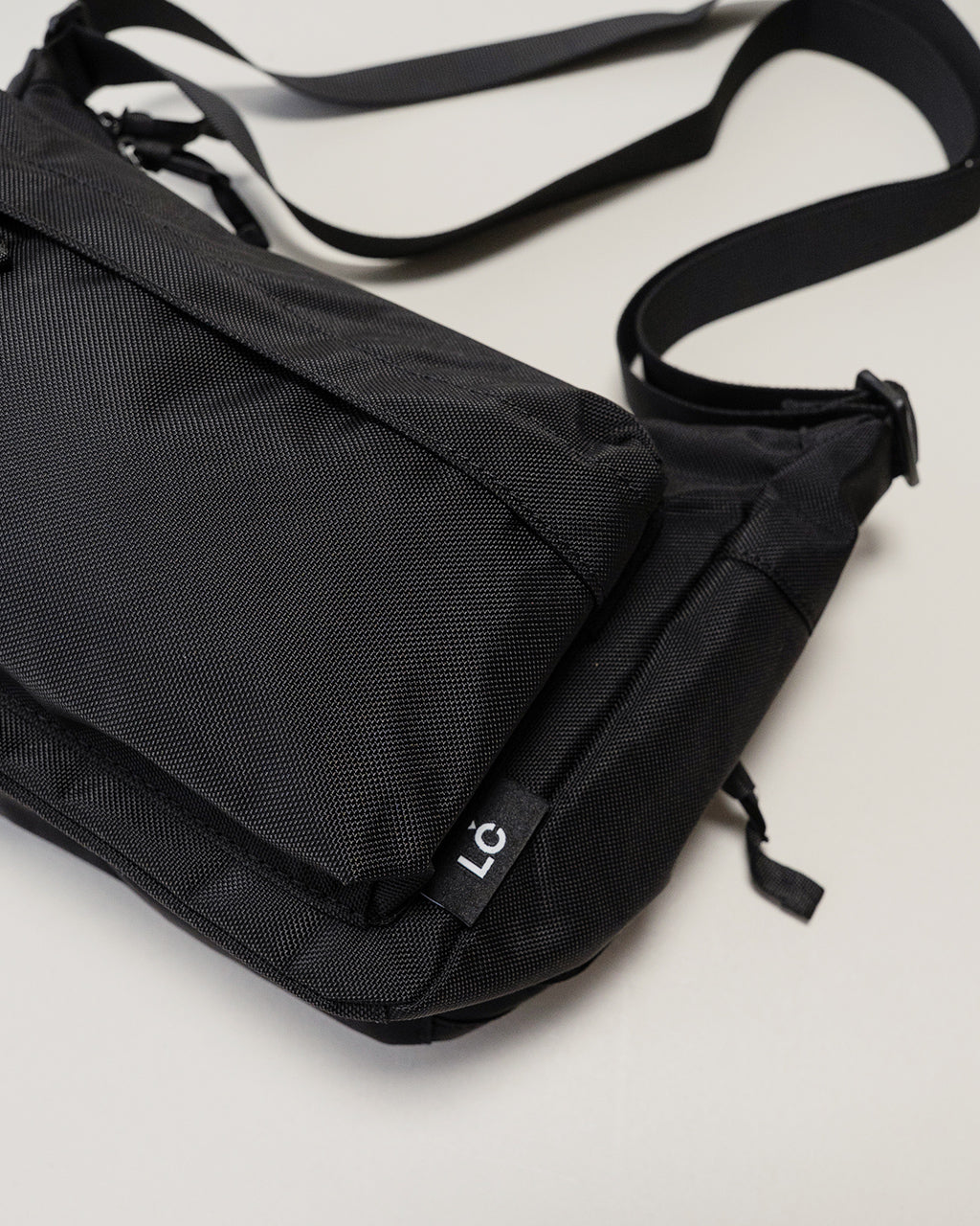 LC by LOWERCASEのブラック系ショルダーバッグ。前後にポケットが見え、丈夫なナイロン素材と黒のストラップが印象的な横からのアングル。ブランドタグが裾付近に見える。