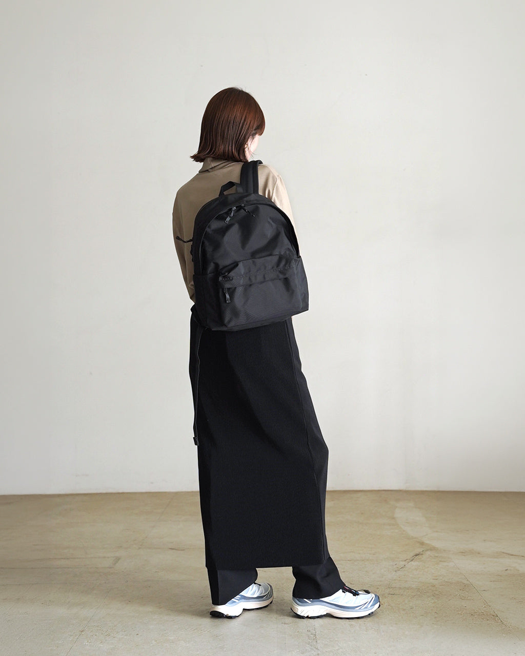黒のバックパックを背負う女性の横姿。シャープなデザインで、前面ポケットとファスナーが目立つバッグの全体像。