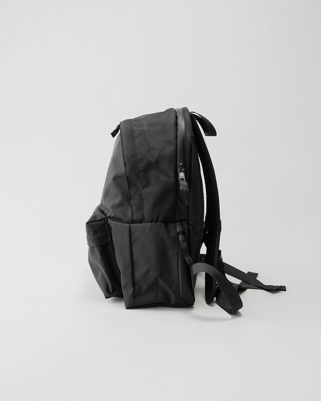 LC by LOWERCASEのブラック系リュック。側面と背面を斜めから捉えたシルエットで、背面ファスナーと肩ストラップ、前ポケットの留具が見える。