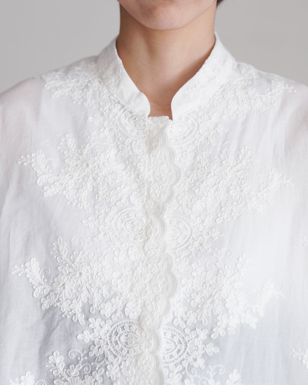 白色のレース刺繍が施されたスタンドカラーのシャツ。繊細な花柄レースが前立てと胸元を美しく装飾しています。
