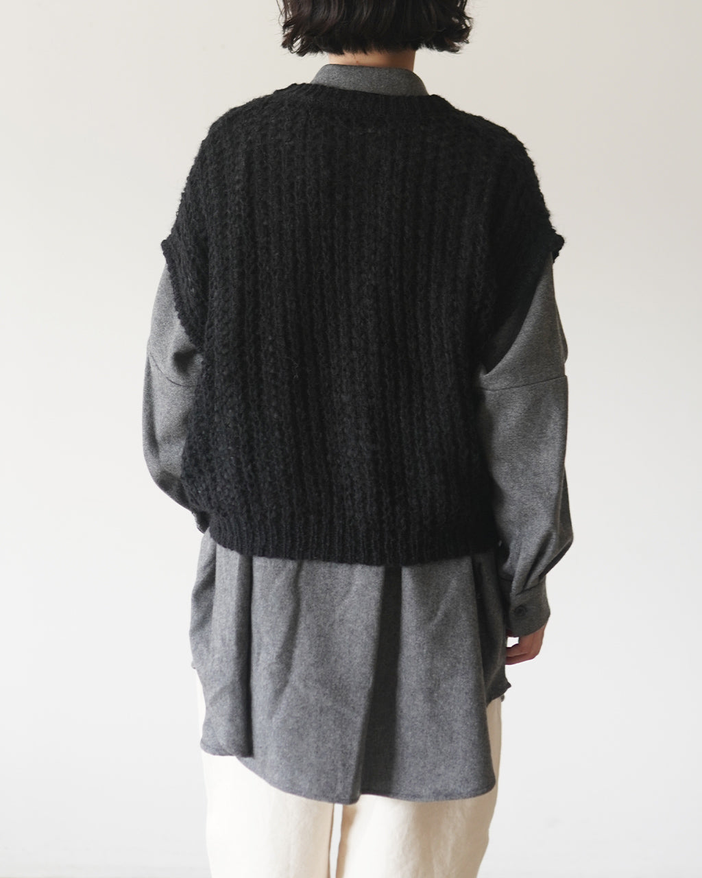 ミズイロインドの hairly cocoon wide vest を背面から捉えた写真。太めのブラックニット編み地が特徴的で、グレーのシャツと重ね着したコーディネートの背中側のシルエットが見える1枚。