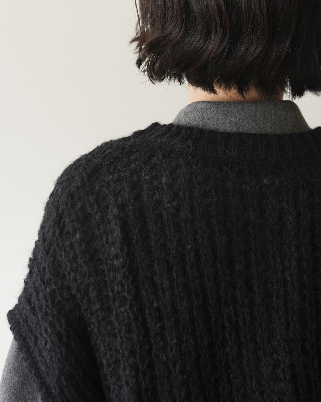 背中側のニットベストを着用した人の首元と肩まわり。黒の編み模様で質感が際立ち、グレーのシャツカラーが覗くディテール。