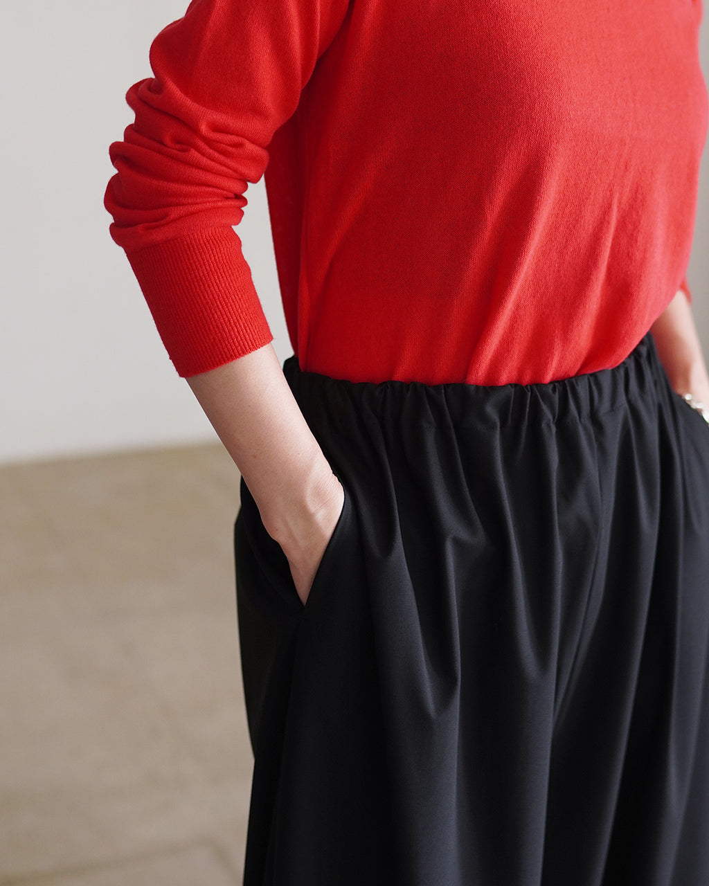 赤いニットと黒のパンツの着用イメージ。ウエストはゴム仕様で、左ポケットに手を入れた状態の近接ショット。