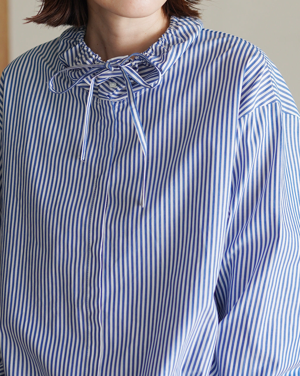 青と白のストライプ柄シャツ、フリル付きネックとリボンタイのディテールが特徴。ややゆったりとしたシルエットで首元のデザインが際立つアップショット