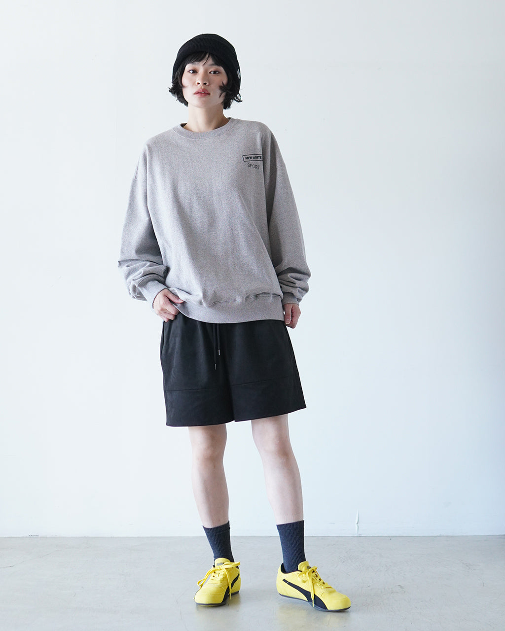 グレーのクルーネックウェアを着用したモデルの全身ショット。ゆったりとしたシルエットと裾リブ、胸元の小さなロゴが特徴。黒のショートパンツと黄色のスニーカーでカジュアルな雰囲気。