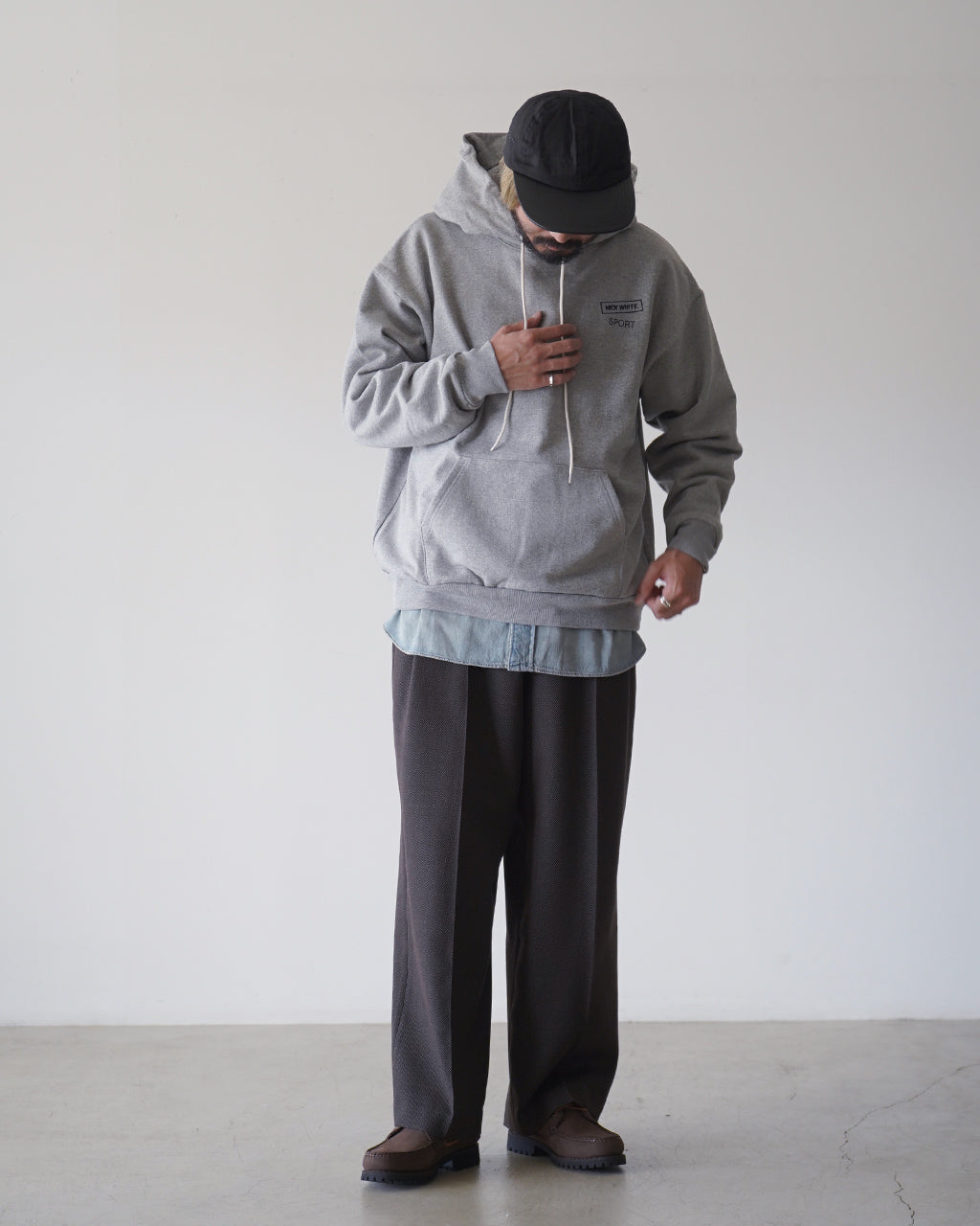 グレーのフード付きスウェットシャツを着た男性の正面ショット。胸元に小さめのロゴがあり、ゆったりとしたシルエットと長めの裾が特徴的です。