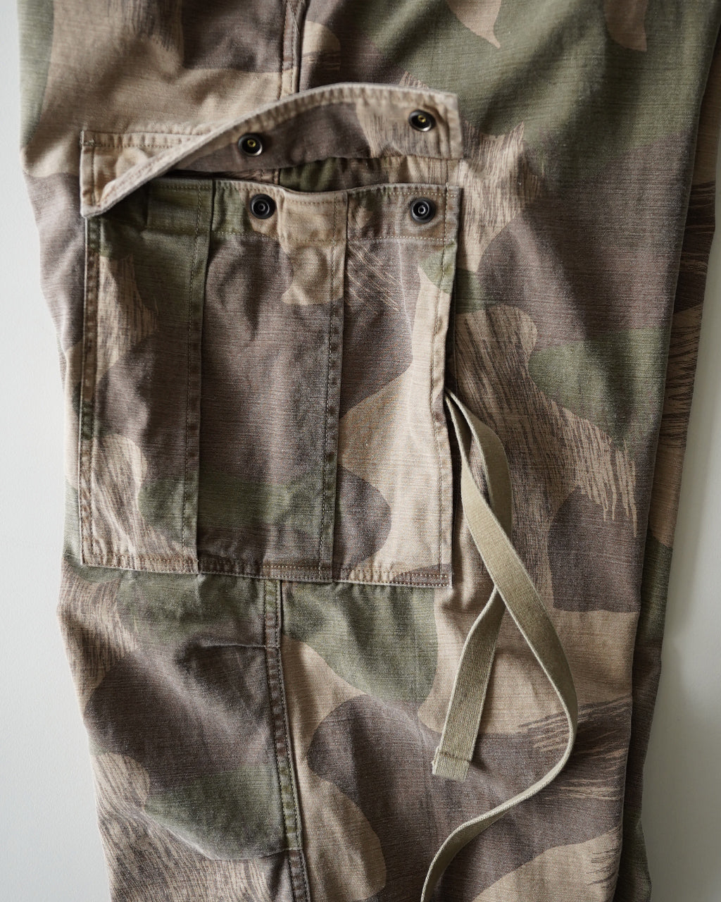 迷彩柄のARMY CARGO PANTの背面と大容量ポケットが映る、米軍風カモフラージュデザインのカーゴパンツ。パネル状のポケットとベルトループが特徴的で、素材の質感が伝わるショット。