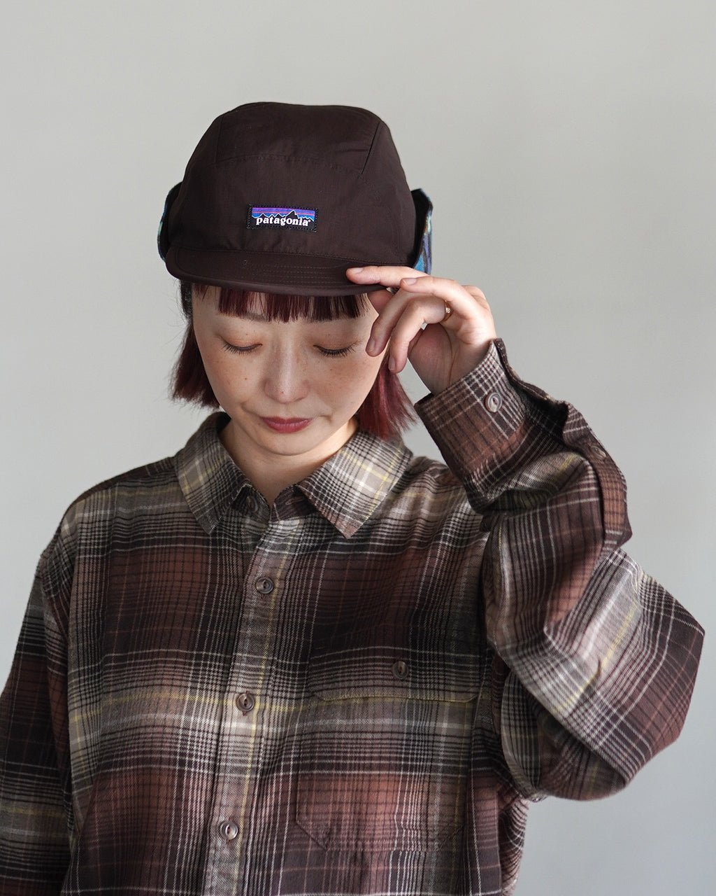 パタゴニアの帽子を被る女性の正面写真。深い茶色のキャップで、前方のロゴパッチが目を引くシンプルなデザイン。チェック柄のシャツを着用している姿を捉えた着用シーン