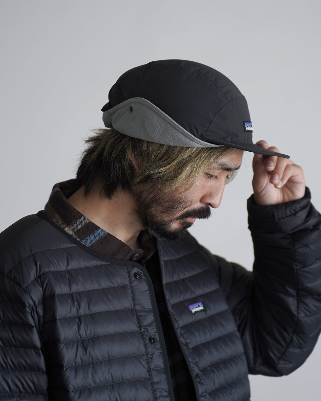 パタゴニアの Isthmus Earflap Cap ブラック系の前方斜めからの全体像。つば広が薄いグレーのイヤーフラップ付きキャップで、ジャケットと同系色のコーディネート。