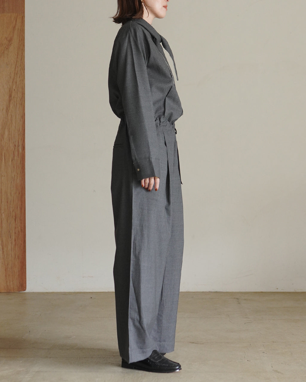 グレーのFine Worsted Jumpsuitを横向きに着用したモデル。袖とウエストのディテールが見え、裾まで落ちるワイドパンツのシルエットが印象的。