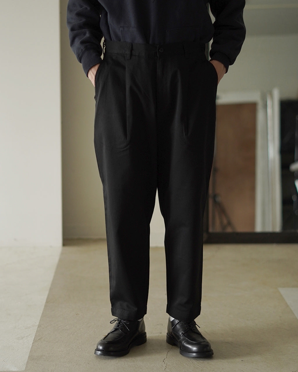 黒系パンツの正面全身ショット。ウエストにタックが入り、裾まで落ちるリラックスシルエットが特徴。モデル着用の黒の上着と合わせたカジュアルな装い。