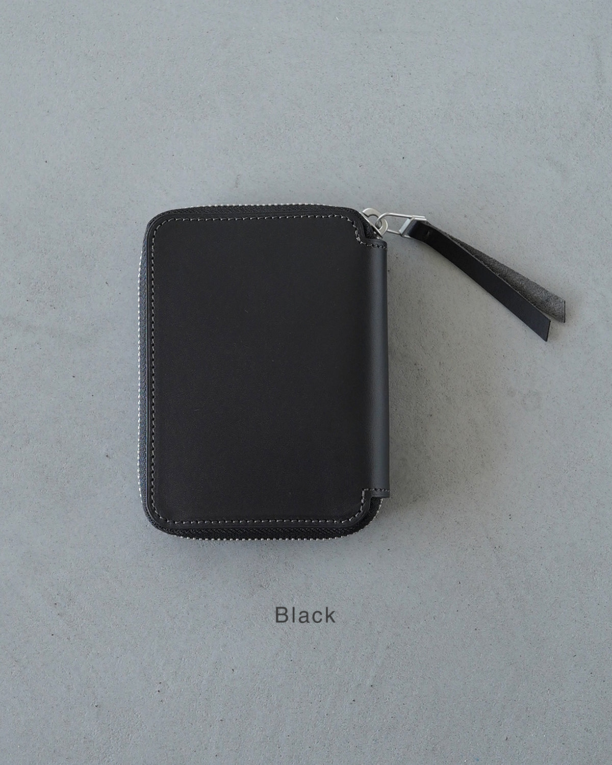 黒のバケッタレザーとホースヌバックが組み合わさった長財布。丸みを帯びたラウンド型のファスナー周りと白いステッチが特徴的で、外観はシンプルなミニマルデザインです。