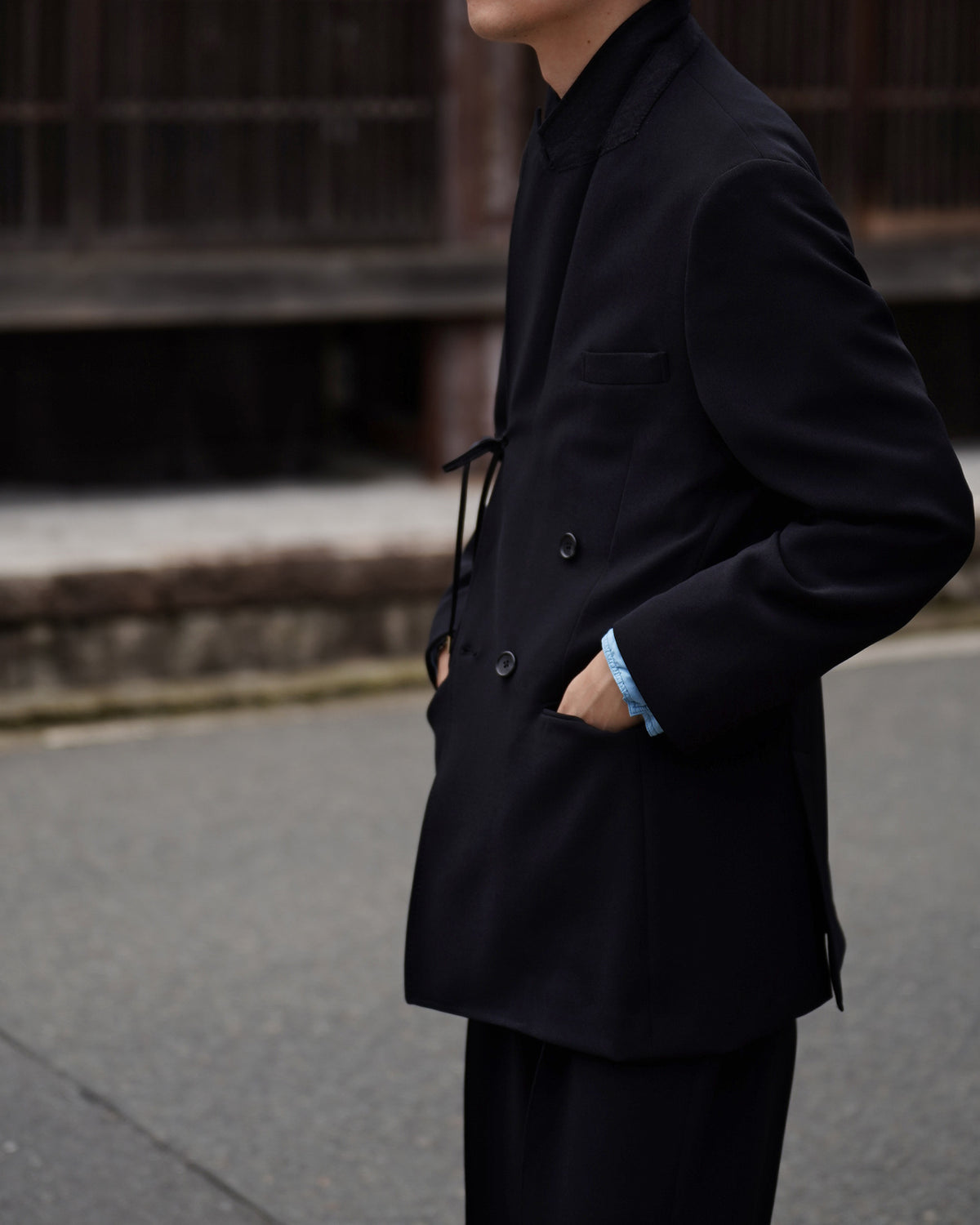 黒いダブルブレストのロングジャケットを着用した男性の横顔と前開き部分を正面寄りに捉えた写真。衿元と肩のラインがすっきり見え、袖口は軽く覗く青みがかったインナーがアクセント。