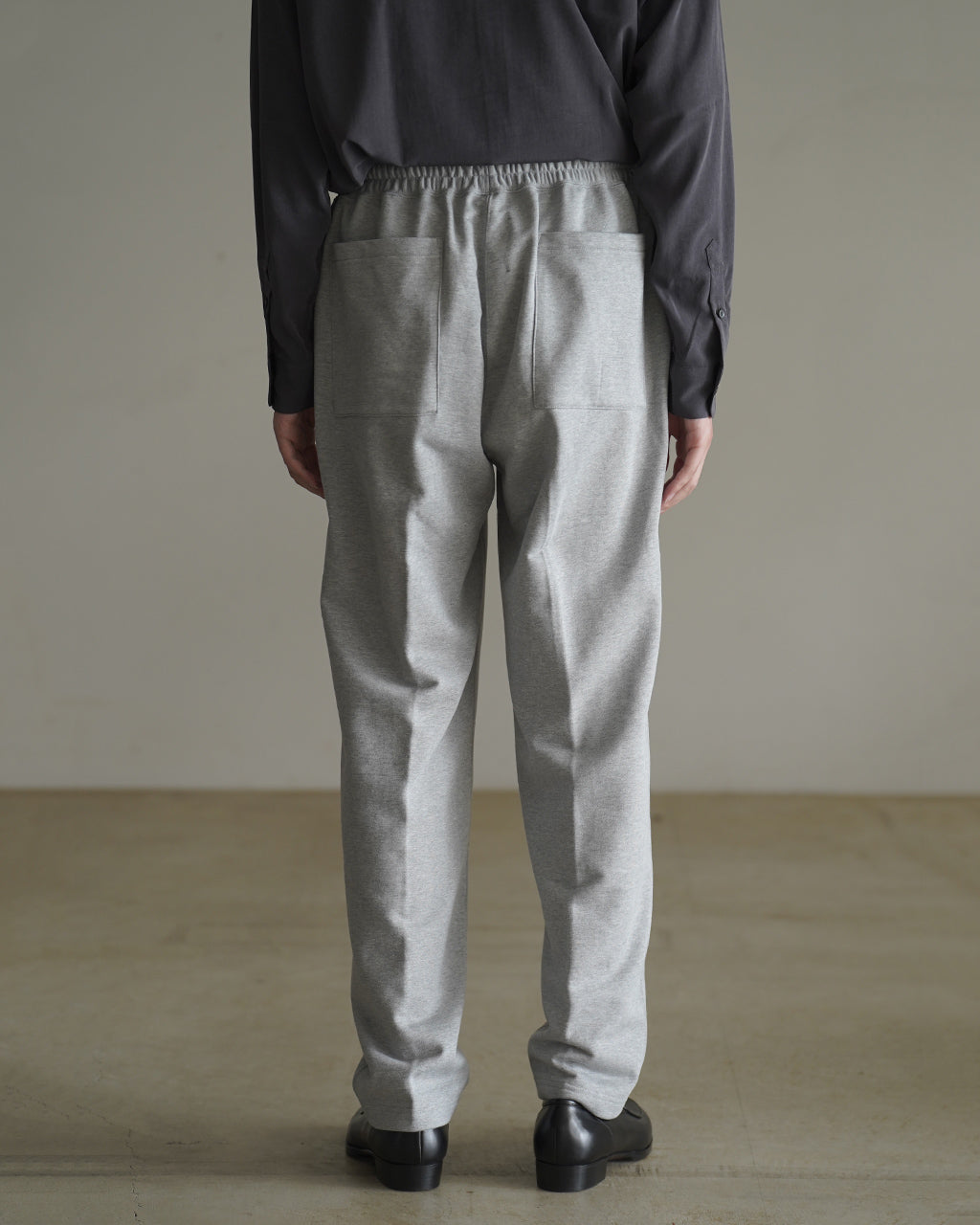 後ろ姿のグレーのEASY SWEATPANTS。ウエストはゴム仕様で、ヒップ周りにゆとりがあり裾に向かってテーパードしたシルエット。背面ポケットが二つ見える。