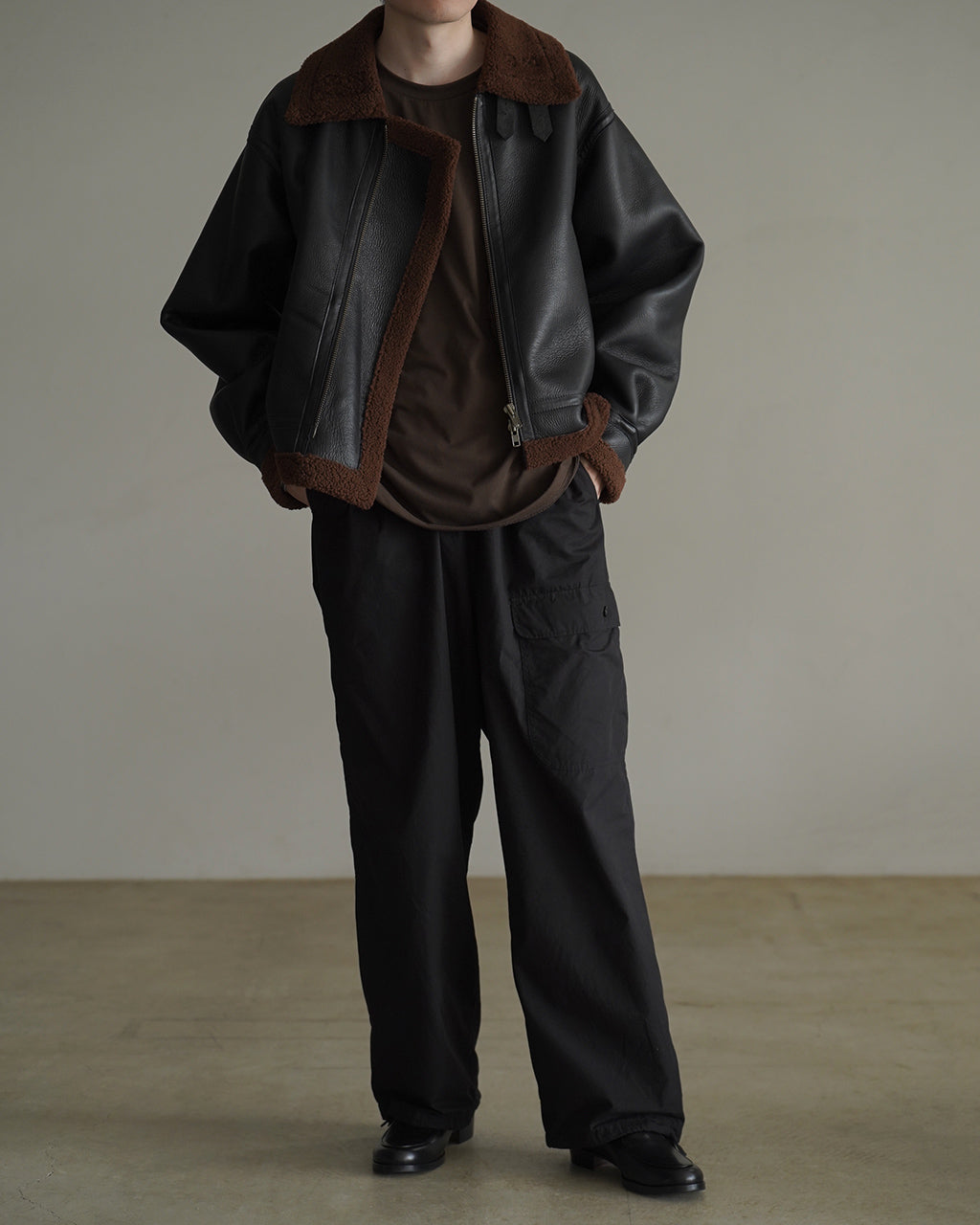ブラックのレザー×ボアのPLEATED AVIATOR JACKETを前面から着用した男性の全身ショット。ベルクロ付きの襟と裾のトリムが特徴的で、袖口と裾に畳まれたプリーツのディテールが見える。