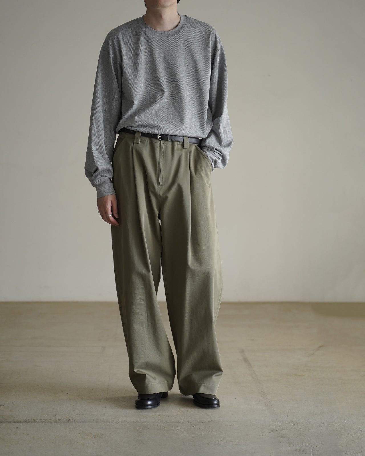 グレーの長袖Tシャツを腰までの高さで着用した男性の前身頃。首元はクルーネック、袖口はリブ付きでゆとりのあるシルエット。下はカーキのワイドパンツと黒の靴でカジュアルながら大人っぽい印象