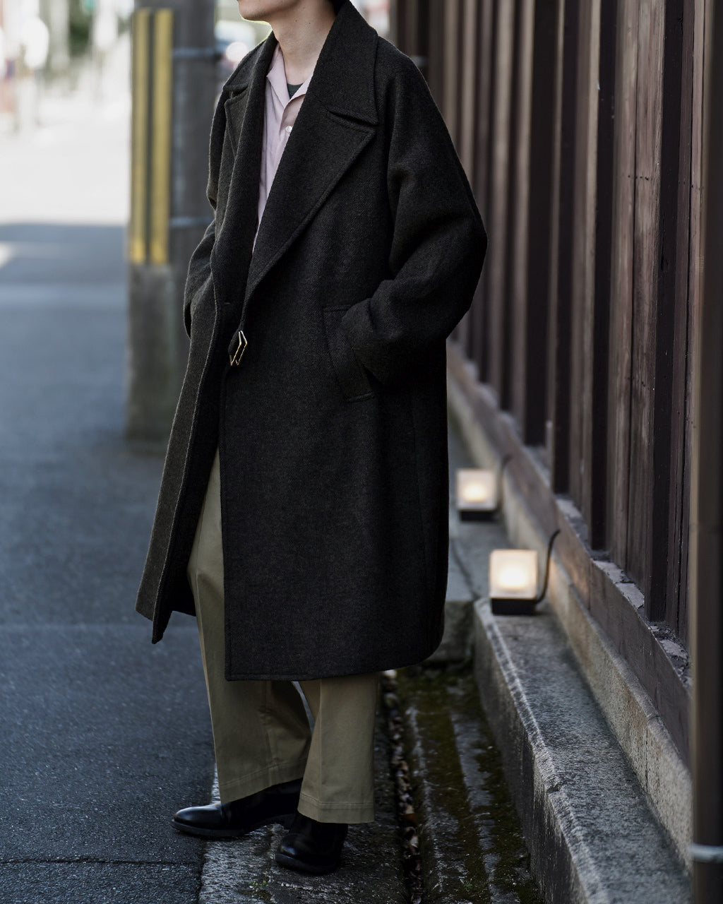 ブラックのウール混コートを着た男性の横姿。肩幅の広いオーバーサイズシルエットで、前を開けた状態に見える。胸元にはベルト留めが見え、裾まで落ちるラインが特徴的。
