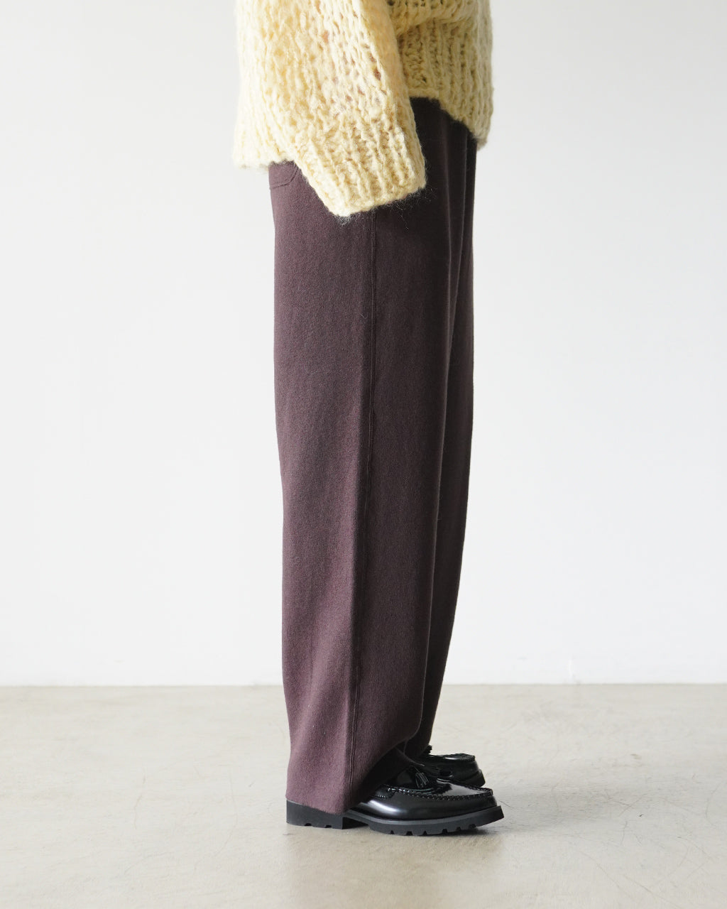 ブランド名RYE TENDERのRoyce Pantsを着用した側面のシルエット。ダークプラム色のパンツとベージュのゆったりニットの組み合わせで、裾が絞られず落ち感のあるデザインが特徴。