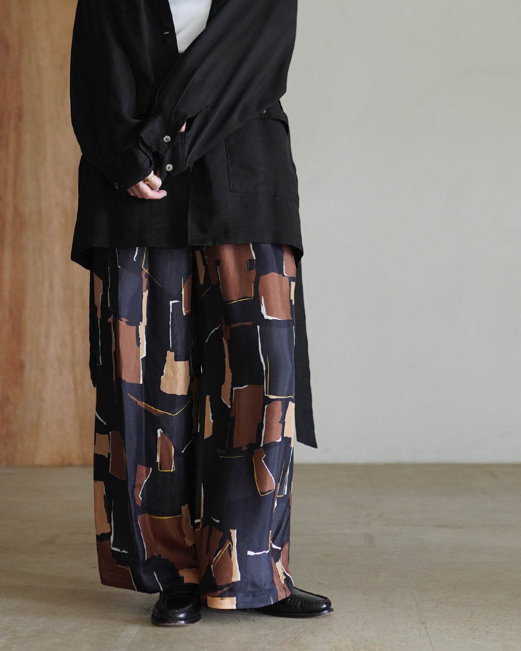 黒のGeometric Painting Pantsが画面左下に見え、シャツ風の黒トップスと組み合わせた着用イメージ。ワイドパンツの柄が全体に広がり、裾までゆとりのあるシルエットが分かる。