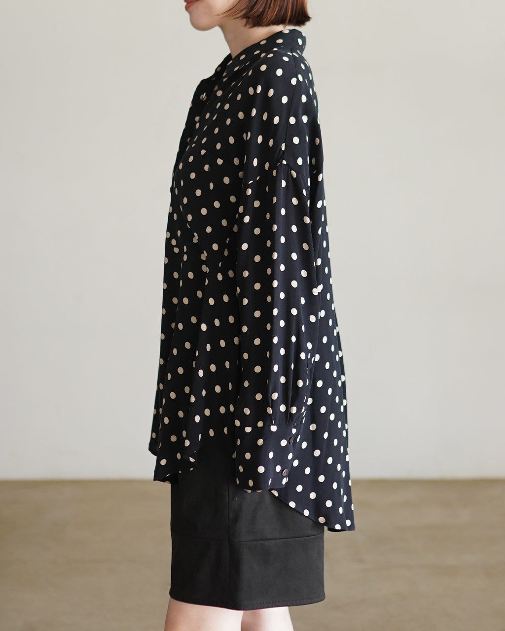 黒地に白のドットが散らばるPOLKA DOT'S SHIRTの横から見たシルエット。ゆったりとしたフォルムと長めのヘムが特徴で、裾はやや後ろ下がりに整えられている。