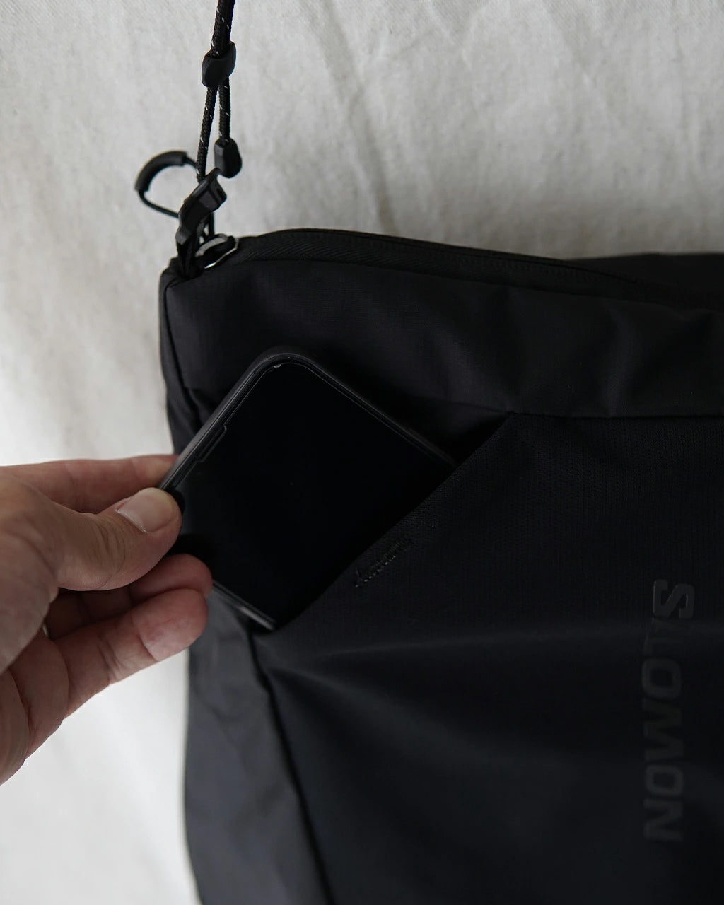 サロモンのACS PACKABLE TOTE BAG、ブラックカラーのナイロン素材をアップで捉えた横持ち写真。スマホをポケットに差し込み、折りたたみ時のコンパクトさが伝わるシーン。