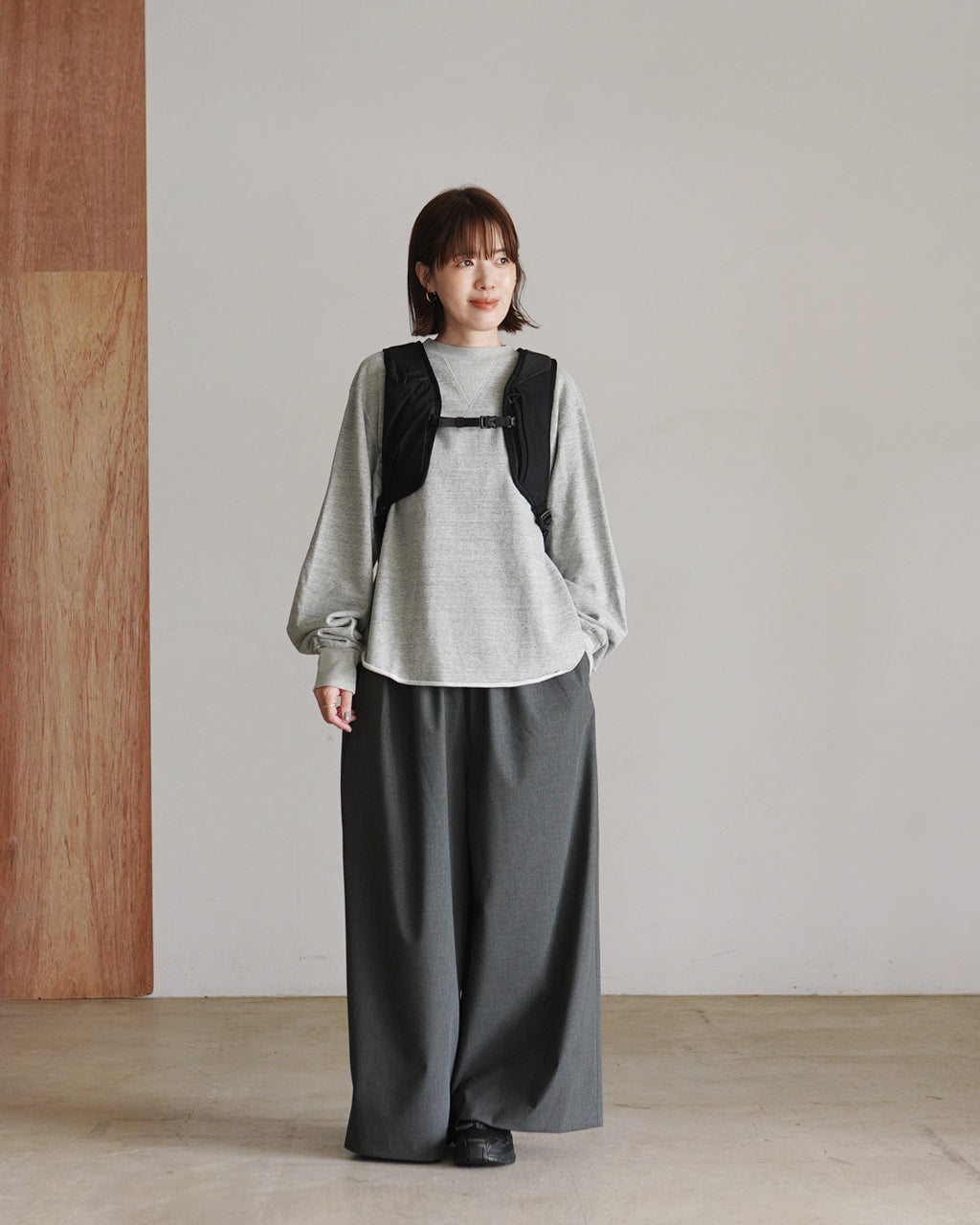 グレーの長袖トップスに黒のバックパックを着用した女性モデルが前方を向くシーン。肩掛けベルトとファスナー付きポケットが特徴的なデザインが目を引く全身ショット